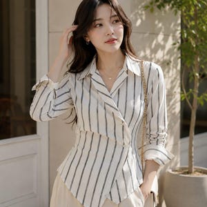 Puede incluir: Una camisa de manga larga con botones, de base blanca con rayas verticales negras. La camisa presenta un cuello de solapa, un cierre frontal cruzado con botones visibles y un dobladillo asim&eacute;trico que termina en punta.