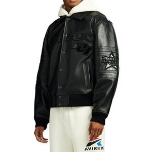 Puede incluir: Chaqueta bomber de cuero negro con el nombre de la marca "AVIREX" en relieve en la parte delantera y la manga. La chaqueta tiene cierre de botones, pu&ntilde;os acanalados y un parche de estrella blanca en la manga. El modelo lleva una sudadera con capucha blanca debajo.