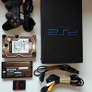 Peut inclure: Une console PlayStation 2 noire avec une manette, un disque dur et une carte mémoire. Des câbles et des connecteurs sont également inclus. La console porte le logo PlayStation 2.