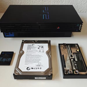 Peut inclure: Console PlayStation 2 noire avec une carte mémoire, un disque dur et un adaptateur réseau. La console porte le logo PS2 et la marque Sony. La carte mémoire est étiquetée "8MB Memory Card" et "FMCB".