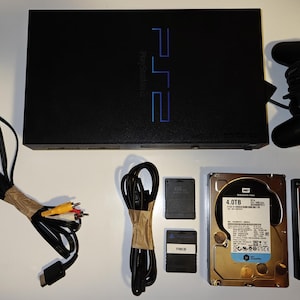 Może przedstawiać: Czarna konsola PlayStation 2 z pasującym kontrolerem, kablami, kartami pamięci i dyskiem twardym 4,0 TB. Konsola ma niebieskie logo PlayStation. Elementy są ułożone na białej powierzchni.