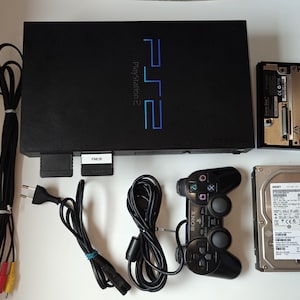 Może przedstawiać: Czarna konsola PlayStation 2 z kontrolerem, kablami i dyskiem twardym. Logo PlayStation 2 jest niebieskie. Dodatkowo karta pamięci i adapter sieciowy.