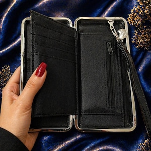 May include: Black wallet with a silver frame, open to show card slots and a zipped compartment. It has a wrist strap with a charm. The wallet is set against a dark blue, textured background.