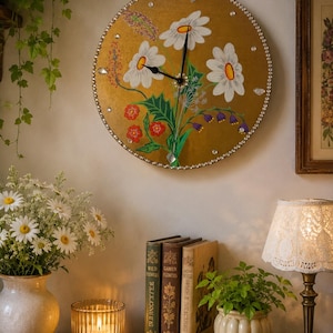 May include: A round wall clock with a gold-toned face featuring a hand-painted floral design of white daisies, small red flowers, and purple bell-shaped blossoms. The clock edge is decorated with a border of small clear crystals and black hands.
