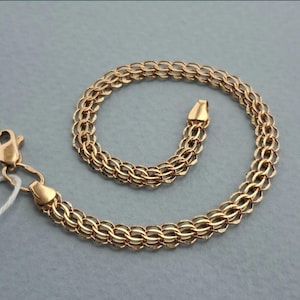 Könnte beinhalten: Ein goldfarbenes Armband mit einem komplexen, mehrschichtigen geflochtenen Kettenglieddesign. Das Armband besteht aus miteinander verbundenen kreisförmigen Metallgliedern, die ein dichtes, strukturiertes Muster bilden. Es enthält einen Karabinerverschluss.