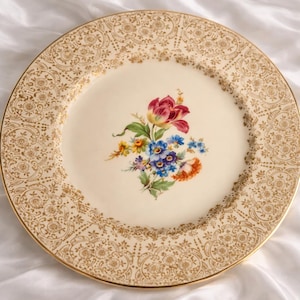 Puede incluir: Un plato vintage de color crema con un borde de filigrana dorado y un ramo floral central. El ramo presenta un tulipán rojo, flores azules, amarillas y naranjas. El plato tiene un borde dorado.
