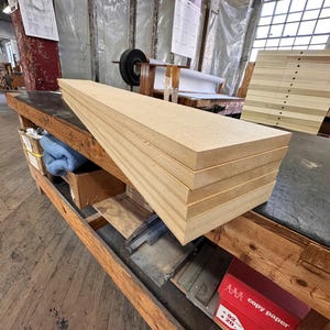 May include: A stack of five rectangular light brown medium-density fiberboard planks with smooth, flat surfaces and straight, clean-cut edges. The boards are layered horizontally, showcasing a uniform, dense, and granular wood-like texture.