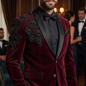 Könnte beinhalten: Ein bordeauxfarbener Samtblazer mit schwarzer Blumenstickerei auf Revers und Schulter. Der Blazer hat ein schwarzes Satinrevers, ein schwarzes Hemd und eine schwarze Fliege. Der Mann trägt eine schwarze Hose.