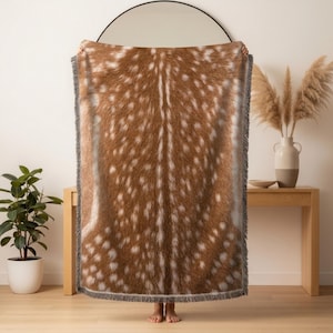 May include: A rectangular woven throw blanket featuring a realistic brown and white spotted deer fur pattern. The blanket has a textured surface with a fringe edge detail along the top and bottom, displaying a dense, soft-looking animal print.