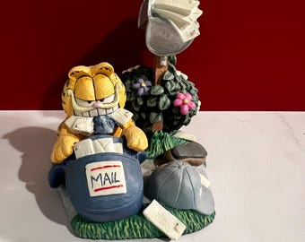 May include: A Garfield figurine with a blue mail carrier uniform, holding a blue mail bag with "MAIL" printed on it. A silver mailbox is behind Garfield, overflowing with mail. A baseball cap and a small white sign are on the grass.