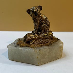 May include: A metallic gold-toned koala figurine perched on a textured log, mounted on a rough-cut, translucent white stone base. The base features the handwritten signature "Rony" in black ink on the left side of the stone surface.