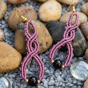 Può includere: Un paio di orecchini in macramè fatti a mano di colore malva, con un design intrecciato, perline nere e ganci dorati. Gli orecchini sono esposti su un letto di piccole rocce e ciottoli. Accessorio unico.