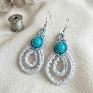 Può includere: Un paio di orecchini in macramè fatti a mano. Ogni orecchino presenta una perla turchese nella parte superiore, circondata da macramè azzurro e un design a goccia in macramè bianco. Ganci per le orecchie color argento.
