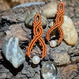 Può includere: Un paio di orecchini in macramè arancione fatti a mano con una perla sul fondo. Gli orecchini hanno un design intrecciato e ganci per le orecchie color argento. Gli orecchini sono esposti su un pezzo di legno.