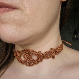 Può includere: Una collana girocollo in macramè marrone con un design intricato e simmetrico. La collana presenta un motivo centrale a forma di fiore ed è legata sul retro con un semplice cordino. Il girocollo è realizzato in un materiale tessuto e strutturato.