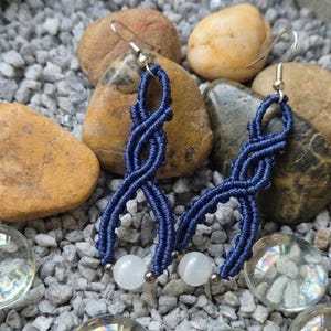 Può includere: Un paio di orecchini in macramè blu fatti a mano con perline bianche e rotonde. Gli orecchini sono esposti su un letto di ciottoli grigi e sono circondati da pietre lisce e marroni e sfere di vetro trasparente.