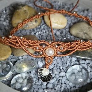 Può includere: Una collana girocollo in macramè color rame con un pendente a perla e perline d'argento. La collana presenta una perla centrale e un ciondolo a forma di conchiglia nera e bianca. La collana è esposta su un letto di pietre grigie.