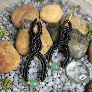 Può includere: Orecchini neri in macramè con ganci e dettagli color oro. Ogni orecchino presenta un design intrecciato con una perla verde e una perla dorata nella parte inferiore. Gli orecchini sono esposti su un letto di ghiaia e pietre grigie.