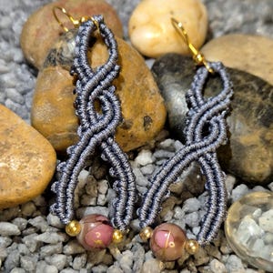 Può includere: Un paio di orecchini macramè fatti a mano. Gli orecchini presentano un design grigio intrecciato a forma di goccia, impreziosito da perline rosa e dorate. Gli orecchini hanno ganci per le orecchie color oro e sono esposti su un letto di rocce.