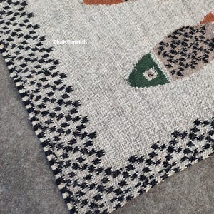 May include: A woven rug featuring a cream-colored base with a black houndstooth border. The rug displays stylized fish motifs in shades of orange, green, and brown with black detailing. The text TheKilimHub is visible on the surface.