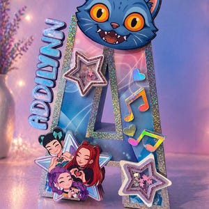 May include: A decorative letter "A" with the name "Addilynn" on the side. The letter is decorated with a blue cat face, stars, music notes, and a group of cartoon girls. The letter is decorated with glitter.