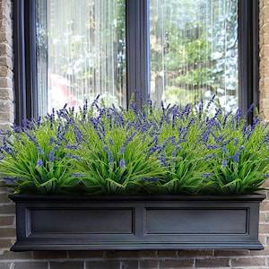 May include: A black window box planter filled with vibrant green and purple faux lavender. The window box is rectangular with a classic design, mounted on a brick wall. The lavender adds a touch of color and natural beauty.