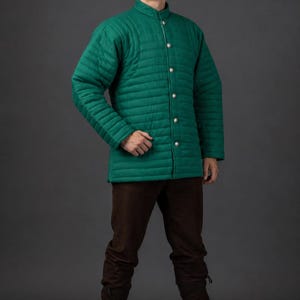 May include: A long-sleeved, emerald green quilted jacket featuring a high collar and a vertical row of silver-toned metal buttons down the front. The garment is paired with dark brown trousers that have a fitted cut and tie closures at the ankles.