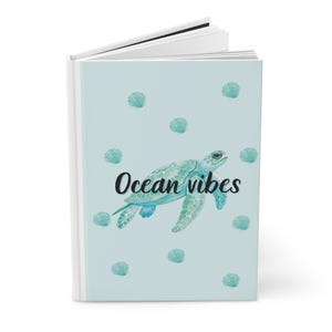May include: A journal with a light blue cover featuring a watercolor sea turtle and scattered seashells. The words "Ocean vibes" are written in black script. The journal has a white spine and pages.