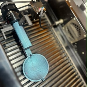May include: A close-up shot of a teal-colored, circular object with a handle, possibly a component of a laser cutting machine. The object is positioned within a metal frame with a series of parallel bars.