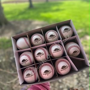 May include: A box of nine white baseballs with red stitching. Each ball has a brown logo and text. The box is held up against a blurred green background. The logo says "Boushier Made".