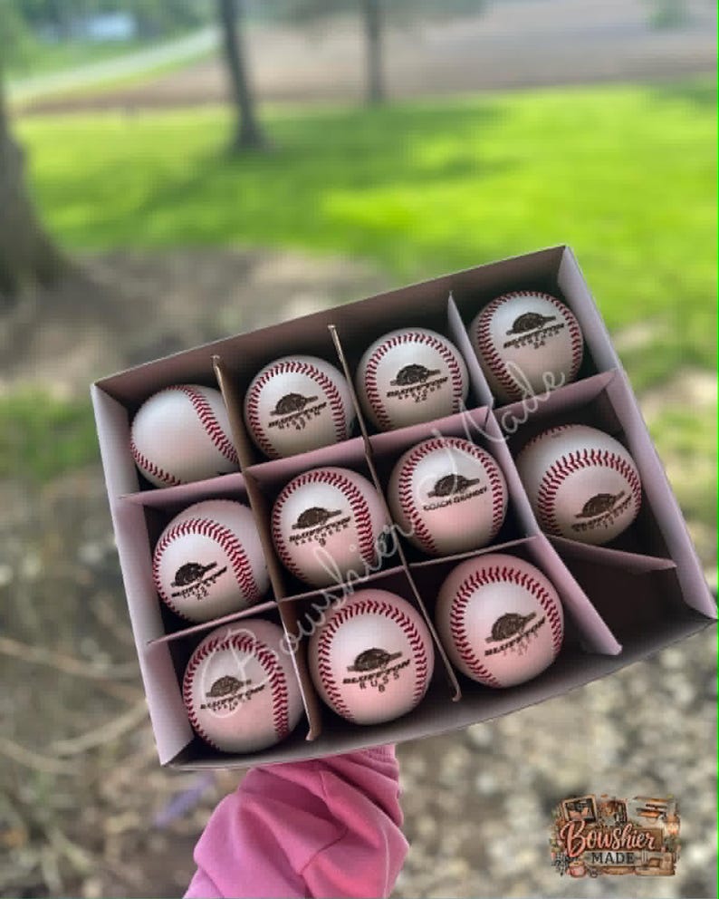 May include: A box of nine white baseballs with red stitching. Each ball has a brown logo and text. The box is held up against a blurred green background. The logo says "Boushier Made".