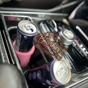 Puede incluir: Interior de coche con dos latas de bebida moradas y un portavasos rosa. Una lata est&aacute; de pie y la otra tumbada. Las latas tienen la parte superior plateada y el cuerpo morado oscuro. Tambi&eacute;n es visible una pegatina decorativa con el texto "Bowshier Made".