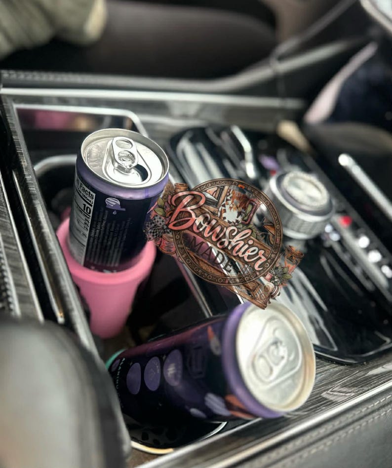 Puede incluir: Interior de coche con dos latas de bebida moradas y un portavasos rosa. Una lata est&aacute; de pie y la otra tumbada. Las latas tienen la parte superior plateada y el cuerpo morado oscuro. Tambi&eacute;n es visible una pegatina decorativa con el texto "Bowshier Made".