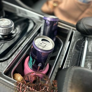Könnte beinhalten: Zwei Aluminiumdosen des Getränks "Cosmic Stardust" in einem Getränkehalter in der Mittelkonsole eines Autos. Eine Dose befindet sich in einem rosa Becherhalter. Die Dosen sind lila mit einem Weltraum-Design. Der Innenraum des Autos ist schwarz.