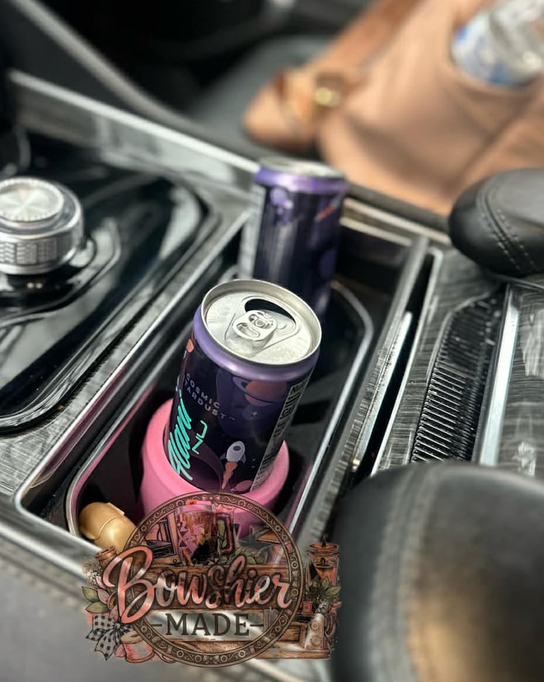 Puede incluir: Dos latas de aluminio de la bebida "Cosmic Stardust" en el portavasos de la consola de un coche. Una lata est&aacute; en un portavasos rosa. Las latas son moradas con un dise&ntilde;o espacial. El interior del coche es negro.