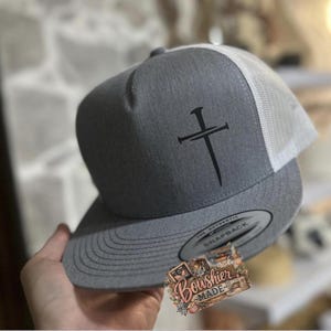 Può includere: Cappellino snapback grigio con un design a croce nera. Ha un retro in rete bianca e una visiera curva. Il testo "Bowshier Made" è su un adesivo sulla visiera.