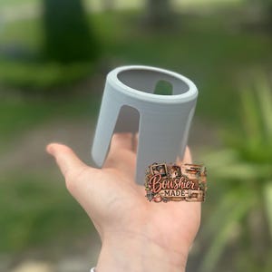 Puede incluir: Un objeto cil&iacute;ndrico gris claro, impreso en 3D, con una parte superior curvada y dos patas arqueadas. El objeto se sostiene en una mano sobre un fondo verde borroso. Es un peque&ntilde;o objeto decorativo.