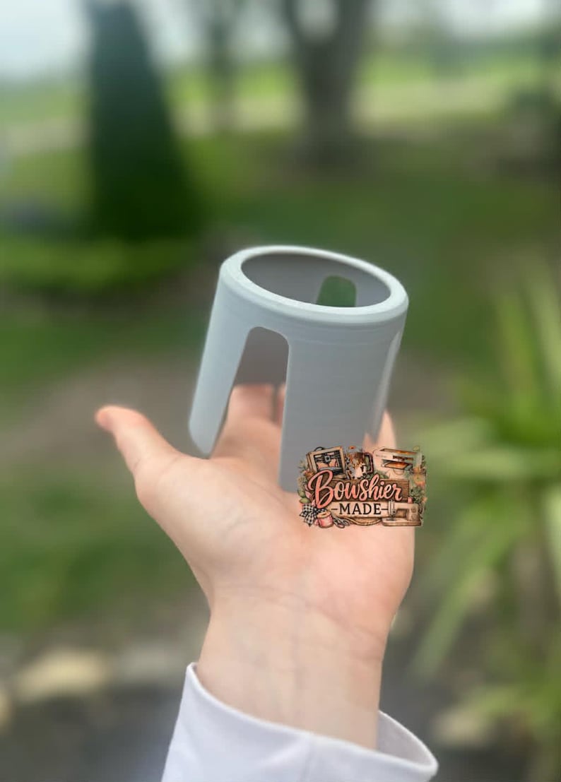 Puede incluir: Un objeto cil&iacute;ndrico gris claro, impreso en 3D, con una parte superior curvada y dos patas arqueadas. El objeto se sostiene en una mano sobre un fondo verde borroso. Es un peque&ntilde;o objeto decorativo.