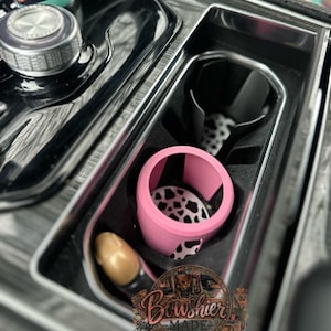 Puede incluir: Un organizador de consola de coche con un portavasos rosa con un dise&ntilde;o de estampado de vaca en blanco y negro. El organizador tambi&eacute;n incluye un art&iacute;culo dorado y un portavasos negro. El logotipo de Bowshier Made es visible.