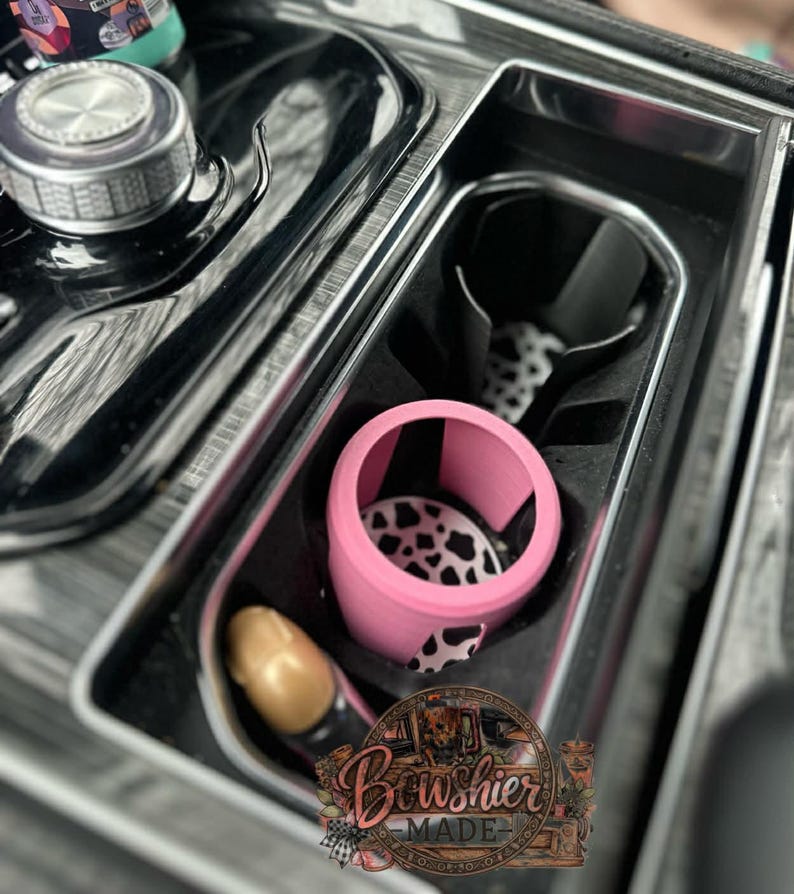 Puede incluir: Un organizador de consola de coche con un portavasos rosa con un dise&ntilde;o de estampado de vaca en blanco y negro. El organizador tambi&eacute;n incluye un art&iacute;culo dorado y un portavasos negro. El logotipo de Bowshier Made es visible.