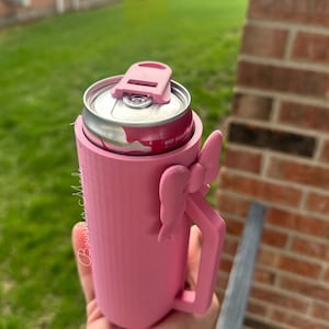 May include: A pink can cooler with a handle and a bow detail. The cooler holds a Red Bull can. The cooler has a lid to keep the can secure. The text "Bourshier Made" is written on the side of the cooler.