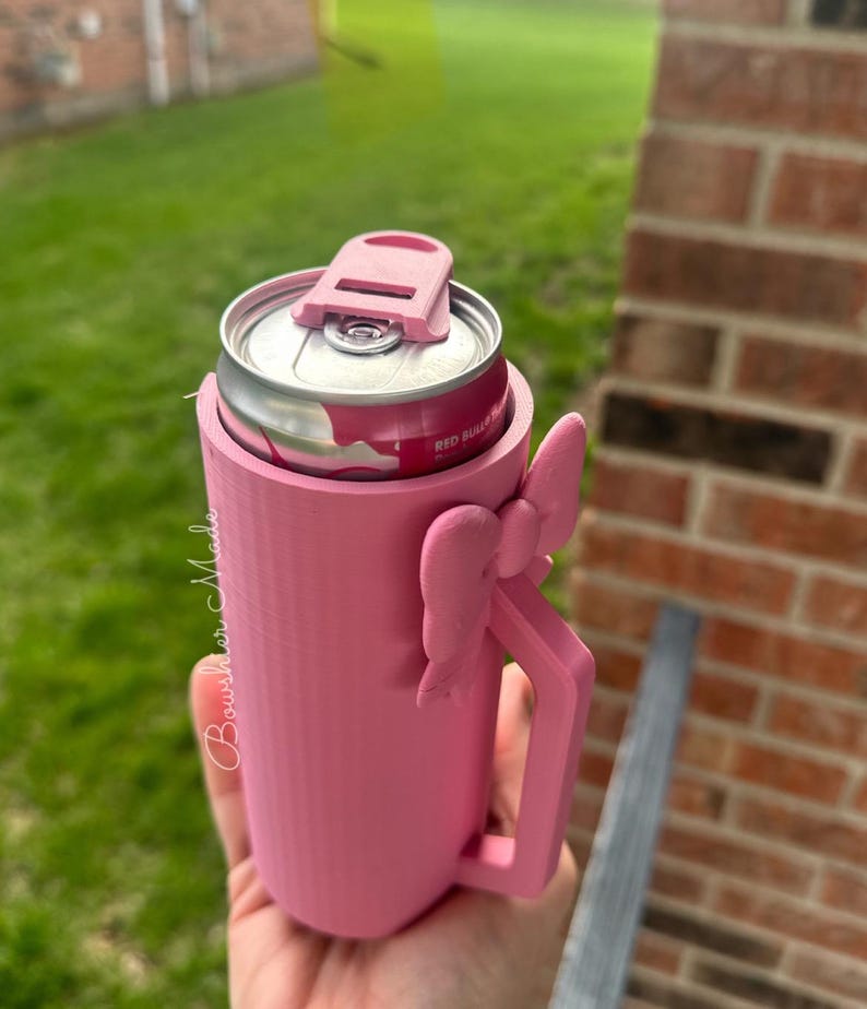 May include: A pink can cooler with a handle and a bow detail. The cooler holds a Red Bull can. The cooler has a lid to keep the can secure. The text "Bourshier Made" is written on the side of the cooler.