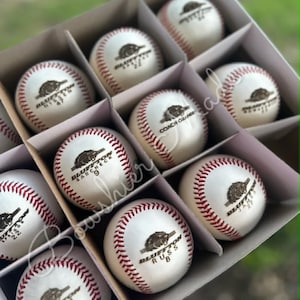May include: A box of baseballs with red stitching and the text "Bluffton" and other text. The baseballs are white with a brown logo. The box is divided into compartments.