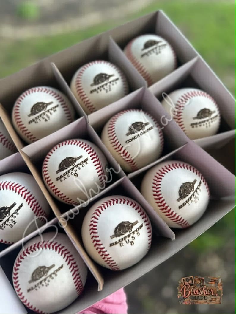 May include: A box of baseballs with red stitching and the text "Bluffton" and other text. The baseballs are white with a brown logo. The box is divided into compartments.