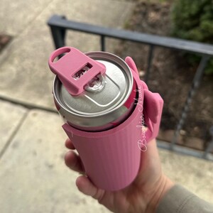 May include: A pink can cooler with a matching lid, designed to keep beverages cold. The cooler is holding a silver can with a pink design. The text "Boughier Made" is visible on the side of the cooler.
