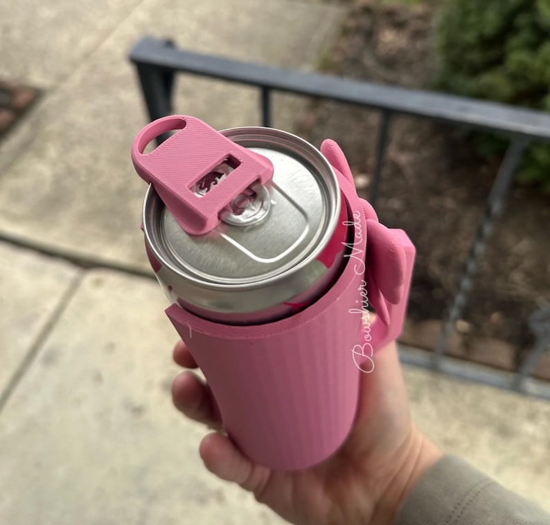 May include: A pink can cooler with a matching lid, designed to keep beverages cold. The cooler is holding a silver can with a pink design. The text "Boughier Made" is visible on the side of the cooler.