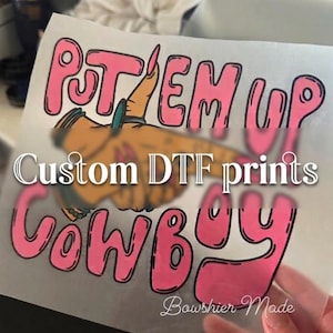 Könnte beinhalten: Eine transparente Folie mit den Worten "PUT EM UP COWBOY" in rosa Buchstaben mit schwarzer Umrandung. Das Bild enthält eine Cartoon-Hand mit einem Ring und eine Cowboy-Grafik. Der Text lautet "Custom DTF prints" und "Bowshier Made".