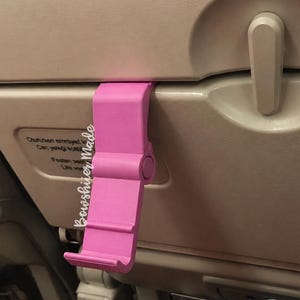 May include: A bright pink phone holder clipped onto an airplane seat tray. The holder is made of plastic and has a slot to hold a phone. The text "Bowshier Made" is visible on the side of the holder.