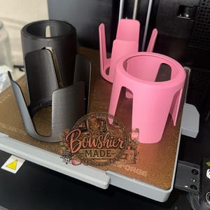 Puede incluir: Una colecci&oacute;n de portavasos impresos en 3D en negro, plateado y rosa. El portavasos rosa es cil&iacute;ndrico con una base ancha y cuatro patas. El portavasos plateado tiene un dise&ntilde;o abierto. La imagen tambi&eacute;n incluye un logotipo que dice "Boushier Made".
