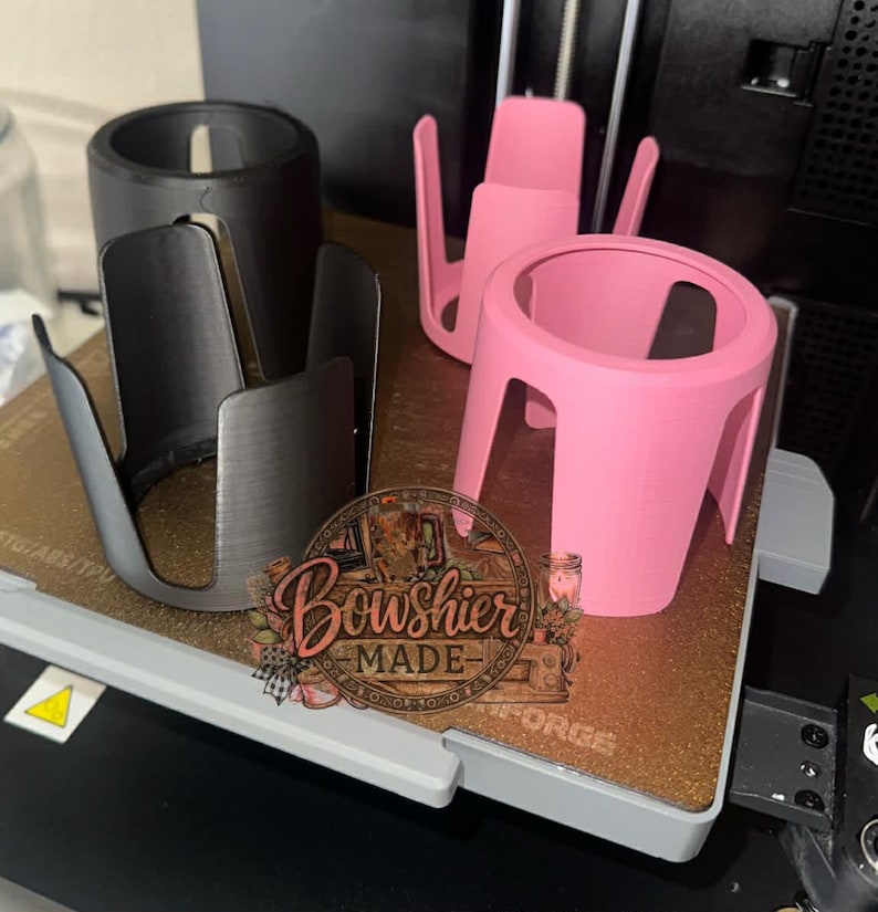 Puede incluir: Una colecci&oacute;n de portavasos impresos en 3D en negro, plateado y rosa. El portavasos rosa es cil&iacute;ndrico con una base ancha y cuatro patas. El portavasos plateado tiene un dise&ntilde;o abierto. La imagen tambi&eacute;n incluye un logotipo que dice "Boushier Made".